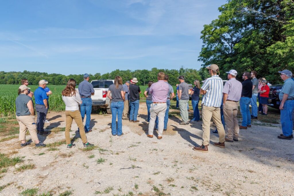 People stand in a farm field watching a person speak. Their backs are to the camera. 