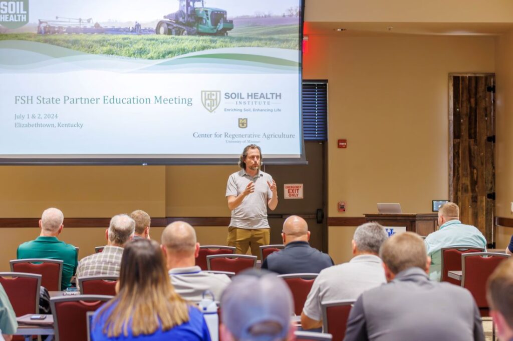 A man speaks to a crowd while standing in front of a screen that reads FSH State Partner Education Meeting