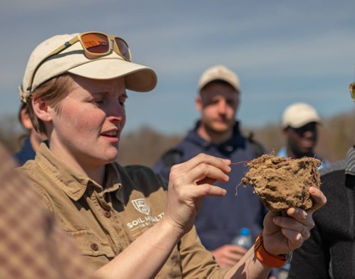 Dianna Bagnall holds a soil clod while speaking to a group of samplers in training. She wears an SHI shirt and a baseball cap.