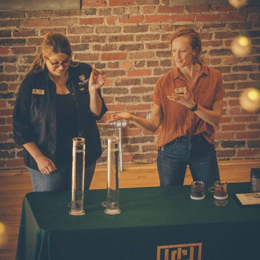 Two women stand in front of a brick wall gesturing to a table where two columns of water stand. One prepares to add a soil clod to the water.