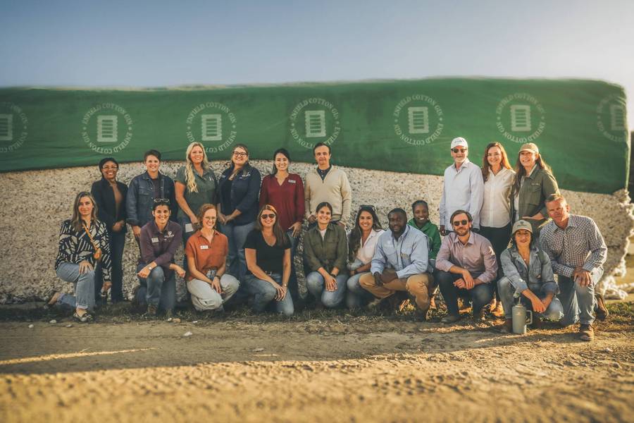 A group photo in front of a large rectangular bale of cotton covered in green fabric that reads Enfield Cotton Gin.