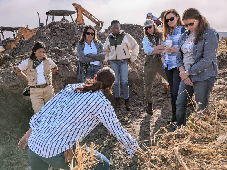 Six women watch a scientist point to the wall of a soil pit. 